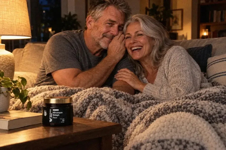 A man of about 62 whispers in his wife's ear on a sofa at night under one blanket, she laughs out loud, the ShilaSource jar on the coffee table beside a paperback