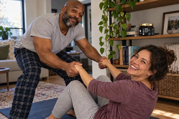 A bald man of about 49 pulls his wife up from a yoga mat by both hands, both laughing, the ShilaSource jar on the shelf behind them